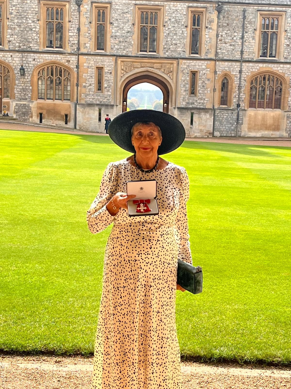 Sue standing in the quadrangle at Windsor Castle in a wide black hat, holding the open box containing her MBE